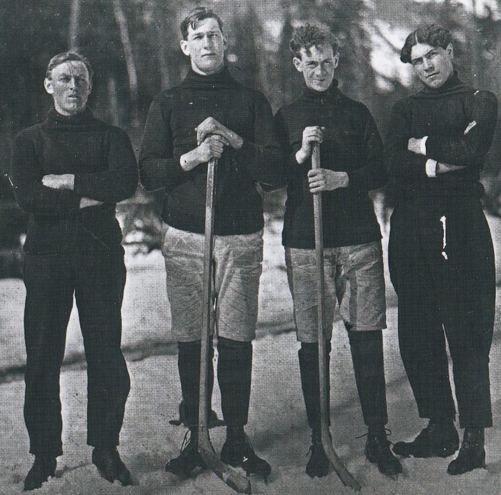 Four young men are standing on ice covered with snow. Two of the men have hockey sticks. All are wearing ice skates and plain sweaters.
