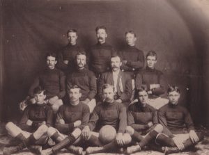 Twelve football players are posing for a picture. The five men in front are sitting with their legs crossed. The man in the middle has a football. All the men are wearing the same sweater except a man in the middle wearing a suit. He appears to be the coach.