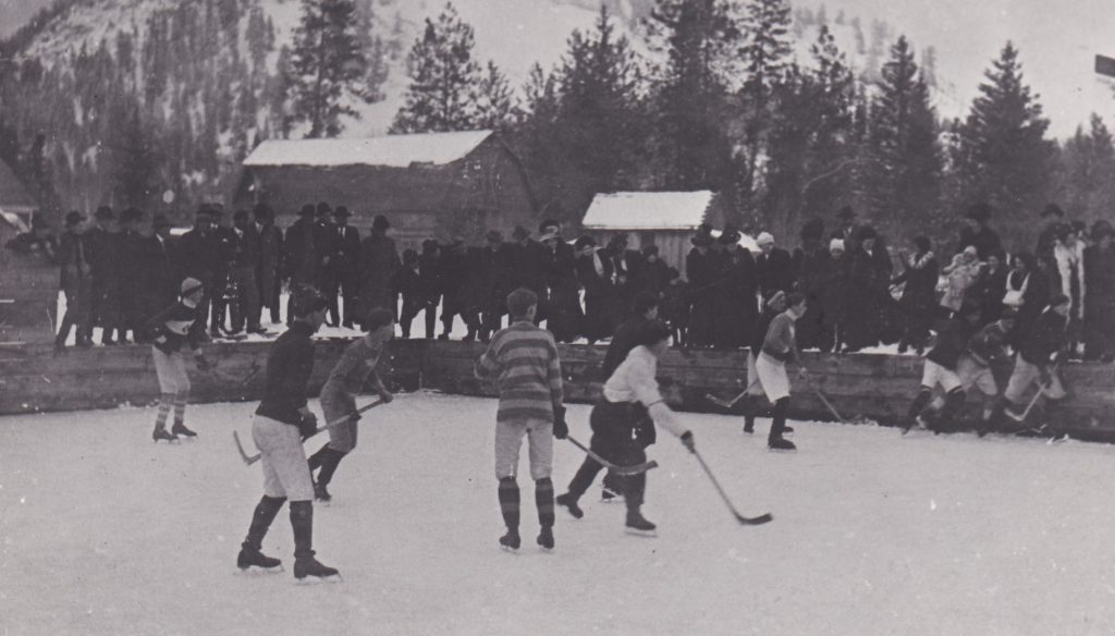 Un groupe de garçons pionniers jouent le hockey sur une patinoire extérieure. Ils sont tous habillés de vêtements différents. Ils ne portent ni casque ni équipement de protection. Une foule observe le jeu.