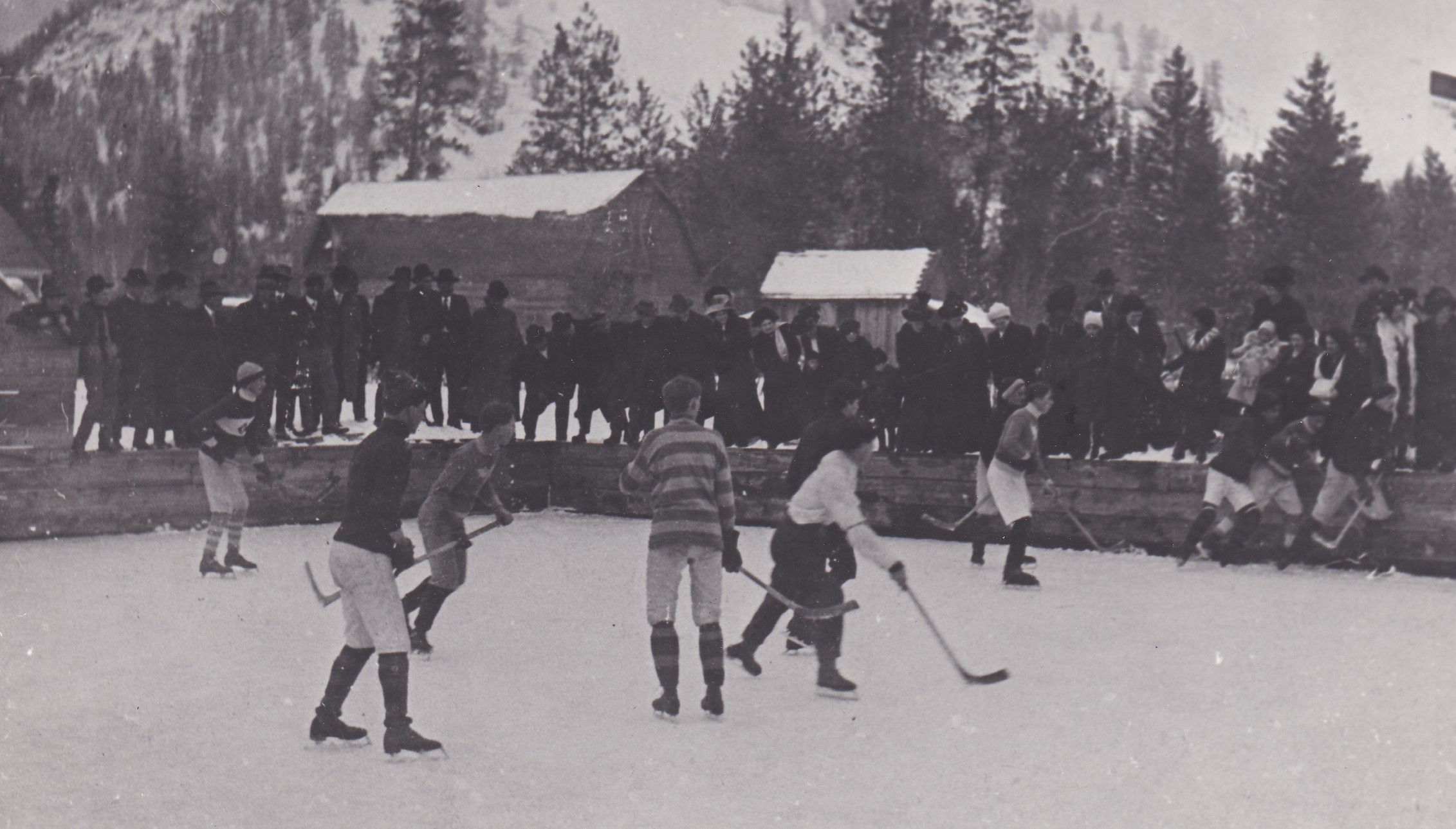 A group of young pioneer boys are playing hockey in an outdoor ice rink. All are dressed differently. None are wearing helmets or any protective gear. A large crowd is watching the game.