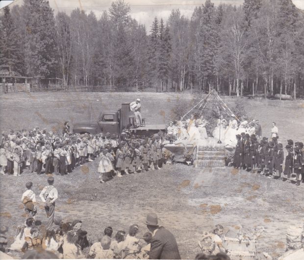 A group of children are assembled in an open field. There is a stage with girls in formal dresses. A truck with a piano is beside the stage. Some of the children are in brownie uniforms.