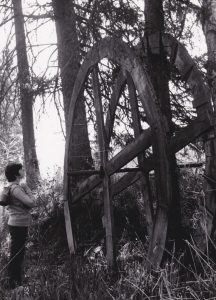 A woman is looking at the remains of a large wooden waterwheel. Tall grass and trees have grown around the waterwheel.