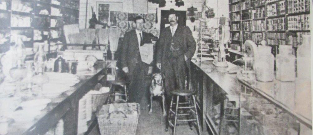 Deux hommes et un chien sont dans un magasin pionnier. Les murs sont couverts par des étagères. Les étagères, les comptoirs et le plancher sont couverts de marchandises. 
