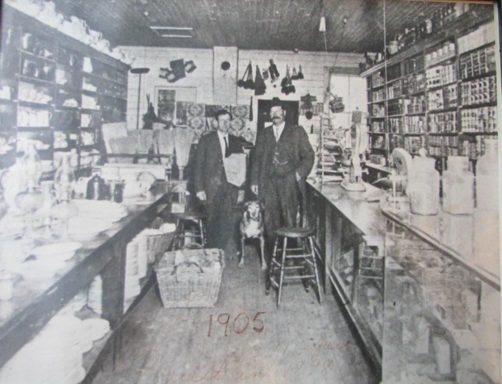 Two men and a dog are standing in a pioneer store. The walls are completely covered with shelves. The shelves, counters and floors are filled with merchandise.