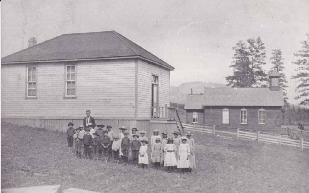 Un groupe d’enfants pionniers et leur professeur sont debout devant une école en bois. Tous les enfants portent des chapeaux. Il y a une église en bois à côté de l’école. 