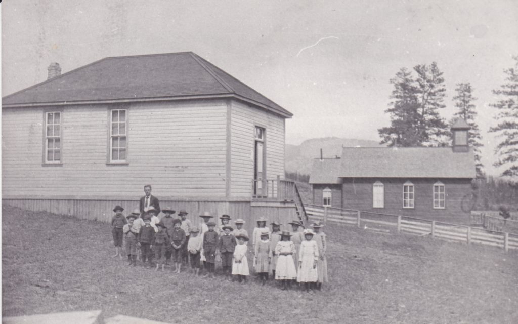 A group of pioneer children and their teacher are standing outside a wooden school. All of the children are wearing hats. Next to the school is a wooden church.