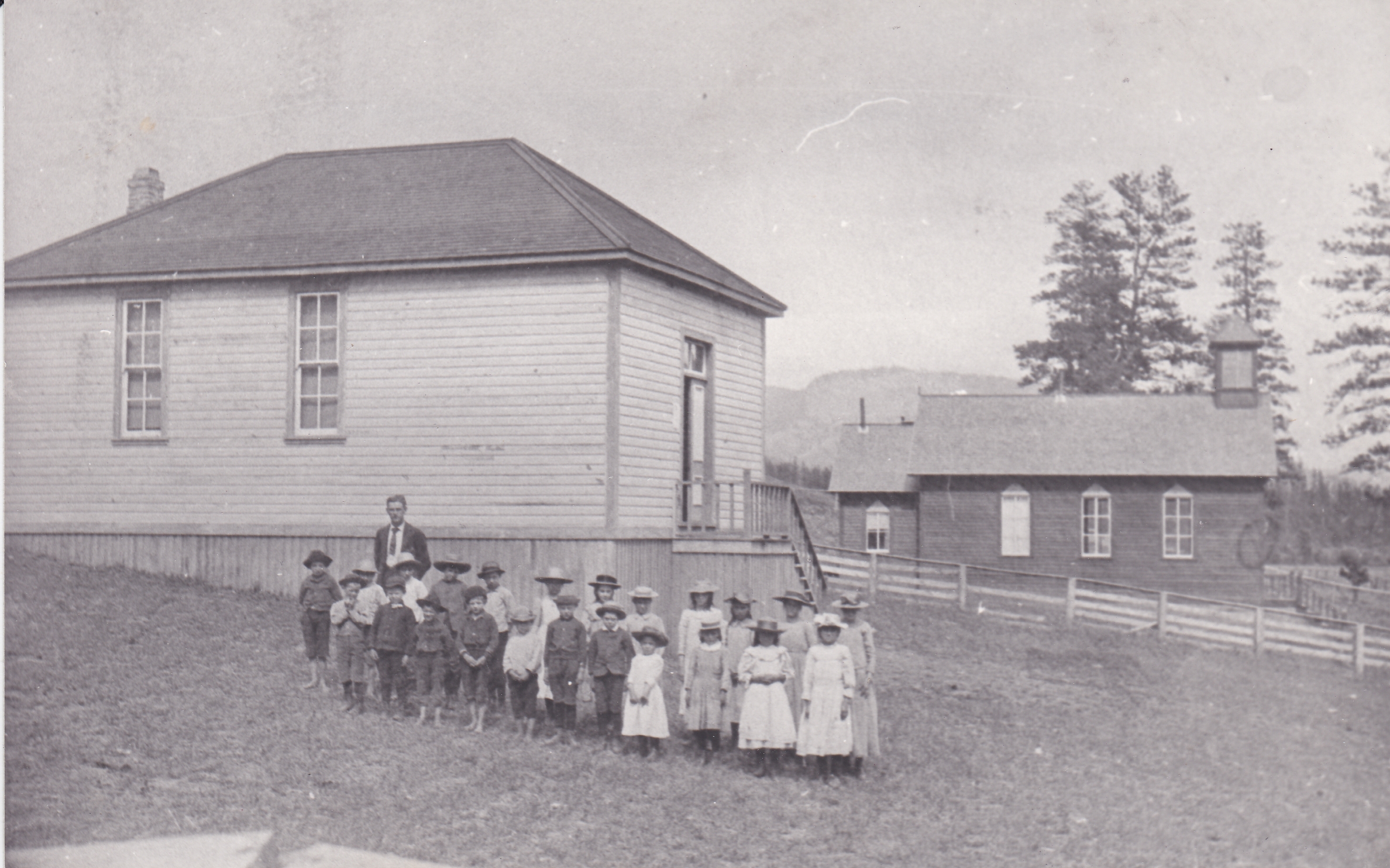 A group of pioneer children and their teacher are standing outside a wooden school. All of the children are wearing hats. Next to the school is a wooden church.