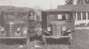 Two school buses are parked on the grass in front of a school. One of the buses is old and square with small windows and narrow tires.