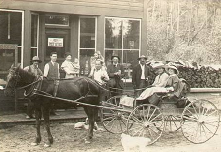 Two women are sitting in a buggy hitched to a horse. The women are wearing old fashioned clothing and hats. A crowd of people are standing on the wooden sidewalk watching them.