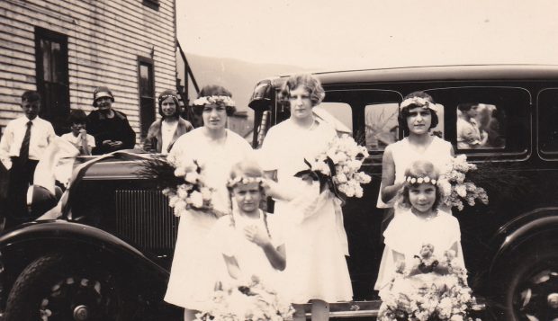 Five girls in white dresses are standing beside an old car. Most have flower garlands in their hair and all are carrying flower bouquets. Two of the girls are young and appear to be flower girls.