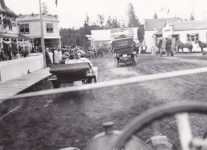 A crowd is standing on wooden sidewalks watching a parade. There are horses and old cars in the parade. The street is dirt and lined with wooden houses and businesses.