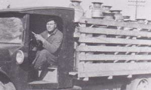 A man wearing a hat and overalls is driving a truck with no doors. The truck has a deck with wooden side walls. The truck is hauling a load of cream cans.