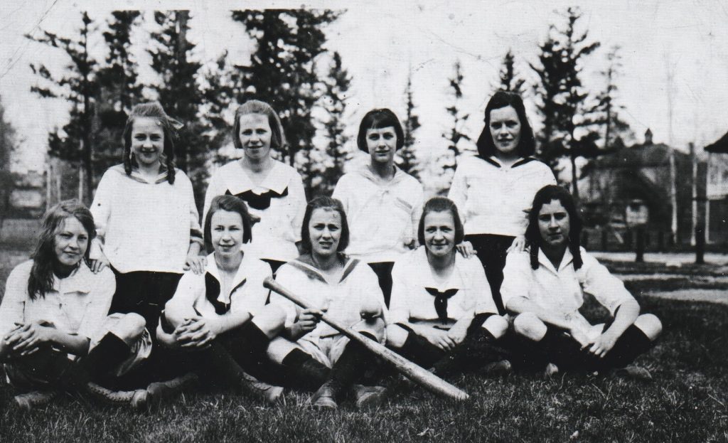A group of female baseball players are sitting on the grass posing for a picture. The players are wearing white blouses, shorts and dark socks. One player is holding a wooden baseball bat and ball.