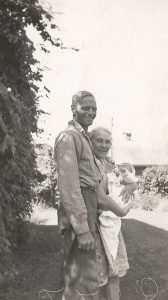 An older man is standing beside an older woman holding a baby. The man is dressed in work clothes and has his left arm on the woman's shoulder. The woman is older and is wearing a cotton print dress that falls just below her knee. The baby is swaddled in a light blanket and has a bonnet on its head.