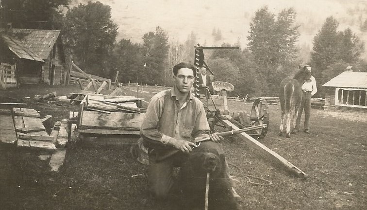 A young man is holding a rifle in a barn yard. He is resting on his right knee behind a black bear. The bear has been shot and its head is propped up with a stick. There are 2 other men standing in the background. One is holding a horse.