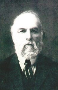 An older pioneer man with a white beard and mustache faces the camera. He is dressed in a formal suit with a cardboard collar and striped tie.