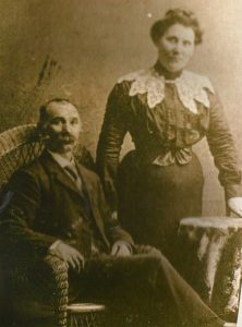 A middle-aged man is seated in a wicker chair. He is balding and has a mustache. He is dressed in a formal suit with a tie. A woman is standing beside him. She is wearing a blouse with a lace overlay, buttoned at the front. Her skirt is long and very narrow at the waist. Her hand is resting on a round table.