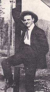 A man in a black suit and ribbon tie is posing on the porch of a house. He has his right hand and right foot on a long metal stake. He is wearing a hat. The house appears to be in a wilderness area as it is surrounded by trees.