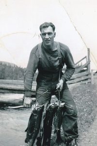 Un jeune homme tient un bâton avec trois saumons percés. L’homme est debout à côté d’une rivière. Il y a un pont avec des planches en bois dans l’arrière-plan.