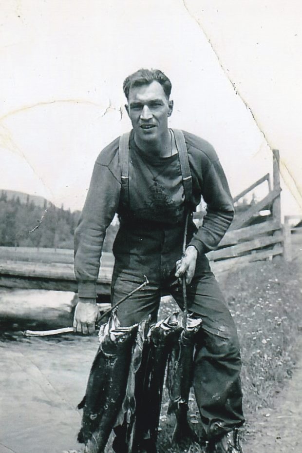 A young man is holding a stick with three large salmon. The man is standing beside a creek. There is bridge with log planks in the background.