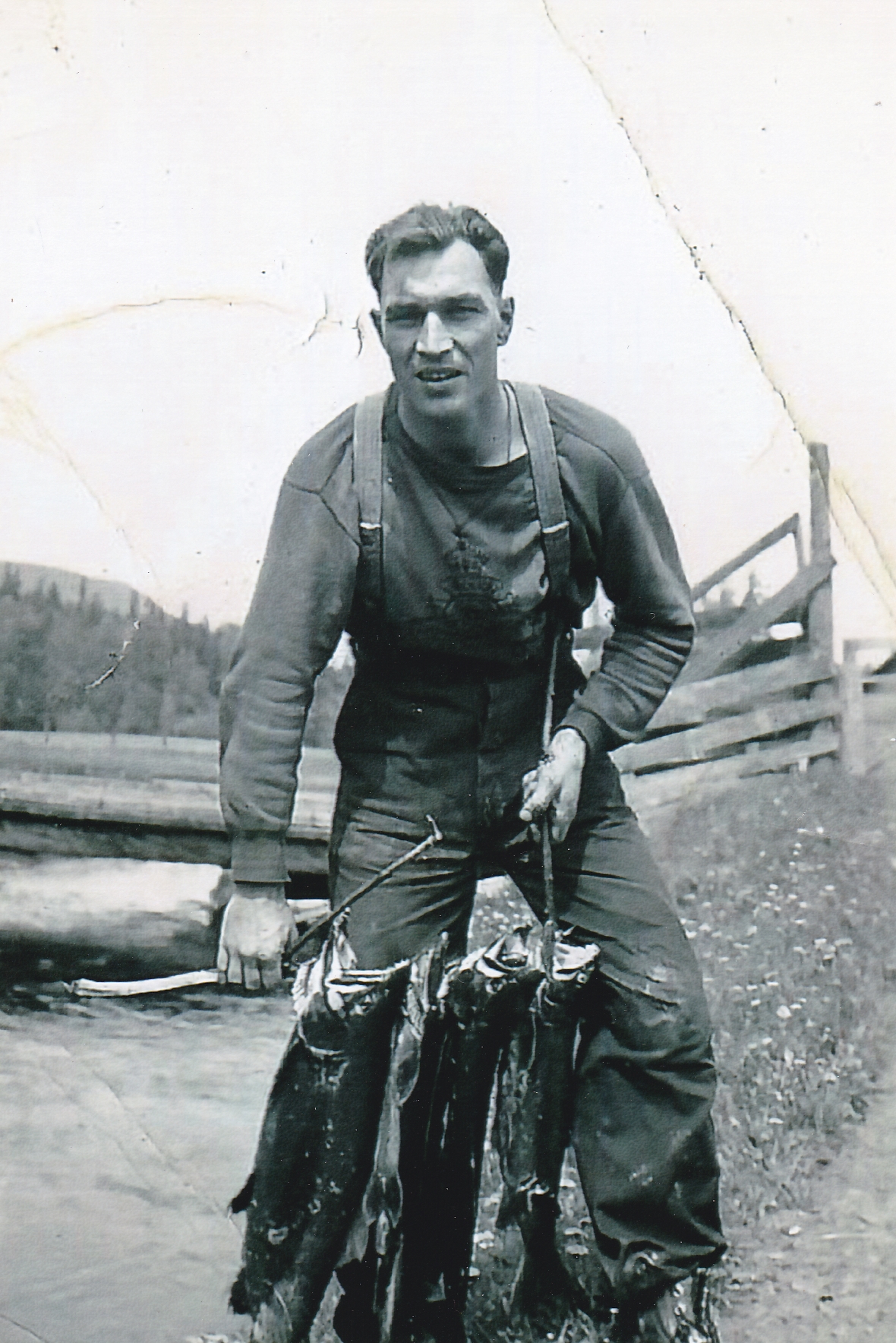A young man is holding a stick with three large salmon. The man is standing beside a creek. There is bridge with log planks in the background.