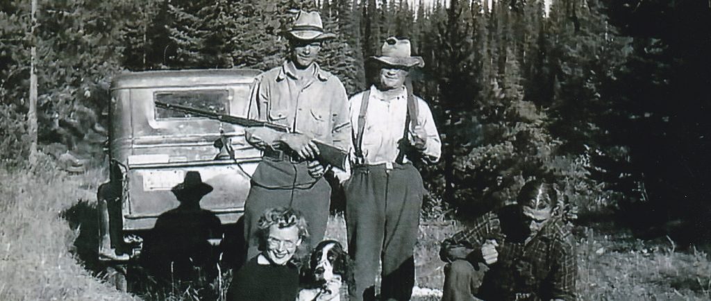 Four people are standing by a truck in the forest. All are holding rifles. Two of the men are standing. A woman is sitting in the foreground petting a dog. Beside the woman are several pheasants that have been shot. A man is crouched in the foreground.