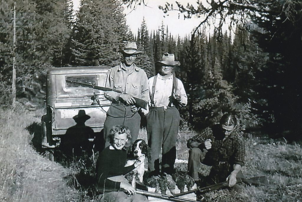 Quatre personnes sont debout derrière un camion dans la forêt. Ils tiennent tous des fusils. Deux des hommes entre eux sont debout. Une femme est assise au premier plan en train de caresser un chien. Il y a des faisans à côté de la femme, abattus par le groupe. Il y a aussi un homme en position accroupie. Il regarde vers le bas.