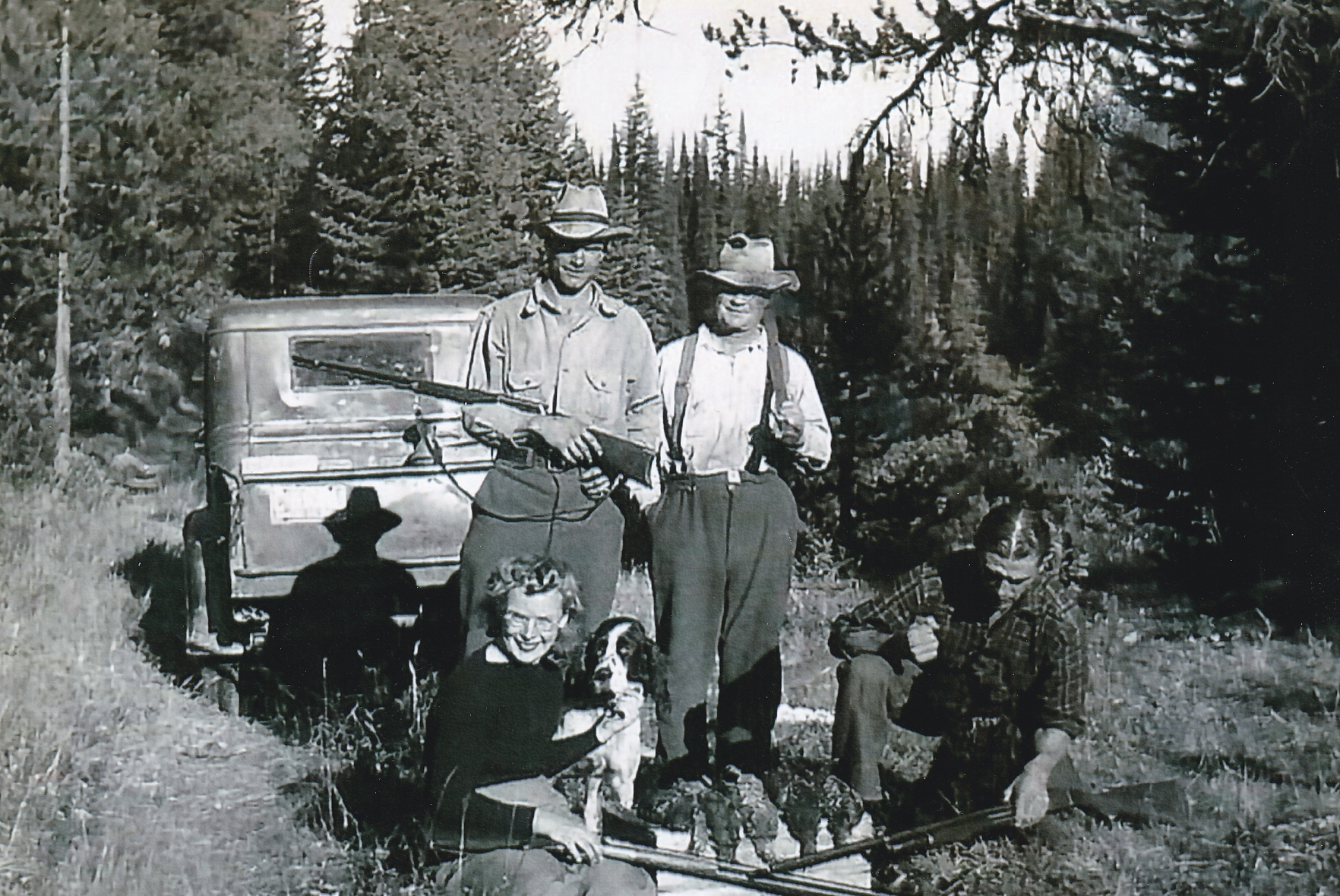 Four people are standing by a truck in the forest. All are holding rifles. Two of the men are standing. A woman is sitting in the foreground petting a dog. Beside the woman are several pheasants that have been shot. A man is crouched in the foreground.