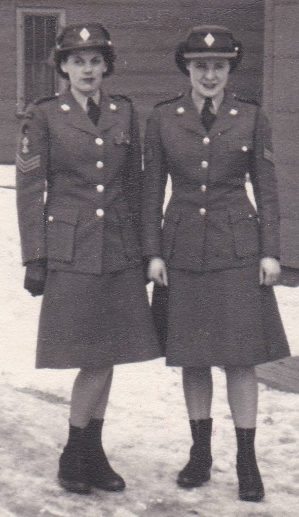 Two women dressed in identical uniforms standing in front of a building. The uniform is a blazer with brass buttons and a skirt just below the knee. Both women are wearing boots. Both women have short hair and are wearing an army hat.
