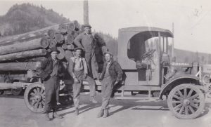 Four men are gathered in front of a pioneer truck loaded with poles. The truck tires are narrow and made of solid rubber. The truck cab is small and open.