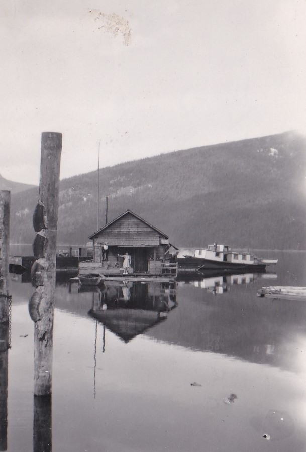 A woman is standing in a rustic wooden house floating on the water. There are large mountains surrounding the water. Two other vessels are tied to the wooden house.