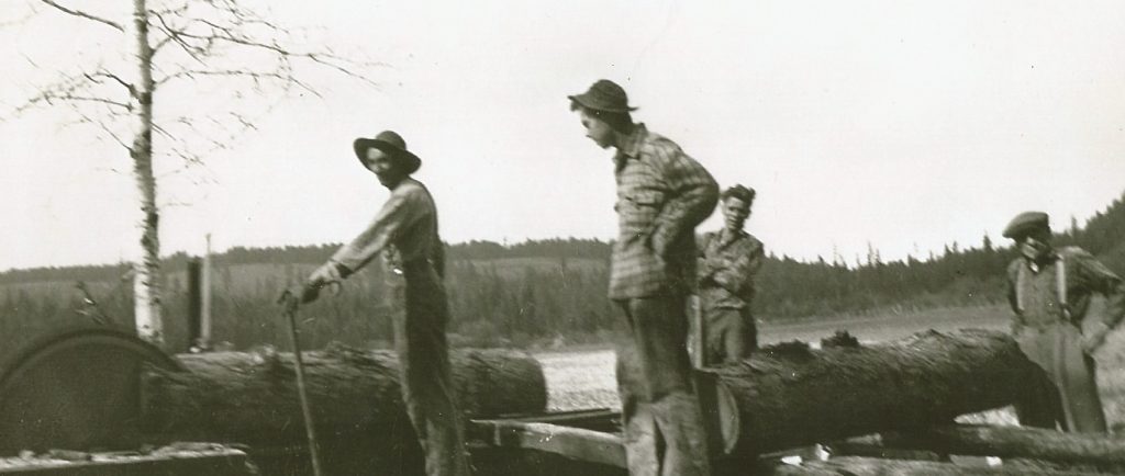 Four men are working beside a primitive sawmill. The saw is cutting a large Larch log and another large log is on the carriage waiting to be cut. The sawmill is in an open field.