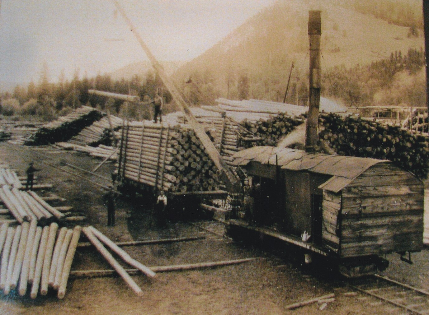 A railcar partly loaded with wooden poles is being loaded by a steam engine. A man is standing on top of the load guiding the poles into place. The steam engine has a long boom with a hook.