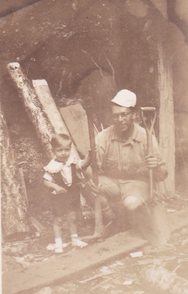 A young boy is standing beside his father who is kneeling in front of the entrance to a mine. The man is holding a shovel and a pick-axe. The man is wearing the little boy's hat.