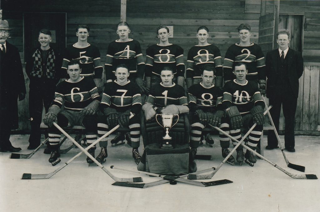 Une équipe d’hockey pionnier tient pose avec un trophée. Il y a deux entraîneurs en costume et un garçon à côté de l’équipe. Les joueurs portent des chandails rayés avec des chiffres. Ils portent aussi des gants, des chaussette rayées et des patins.
