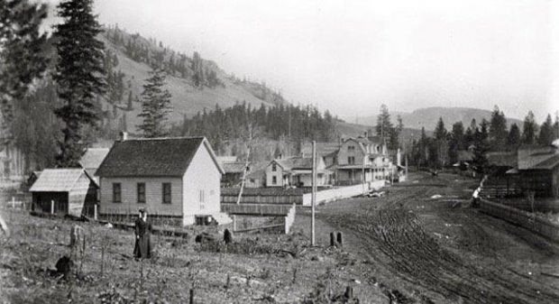 A pioneer woman is standing next to a wooden building. There is a muddy dirt road going past the house. There are several other wooden structures in the background.