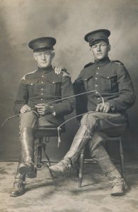 Two WWI soldiers are sitting on chairs. Both are holding riding whips. The younger soldier has his hand on the older soldier’s shoulder.