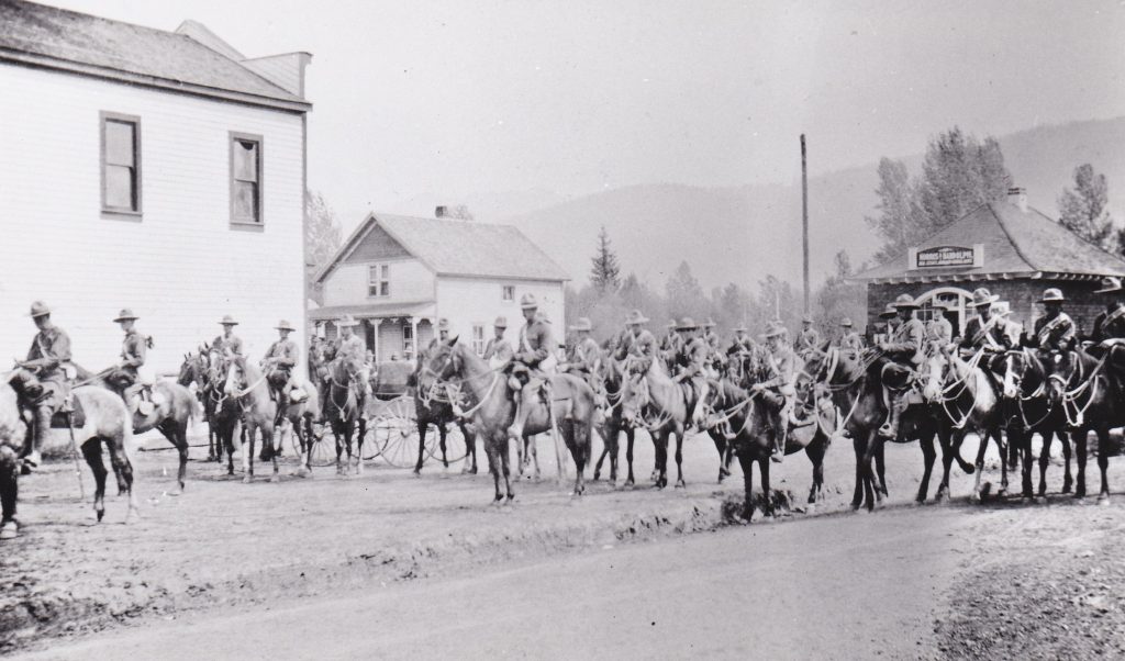 A large group of soldiers are mounted on horseback on a dirt street. There are several wooden buildings in the background.