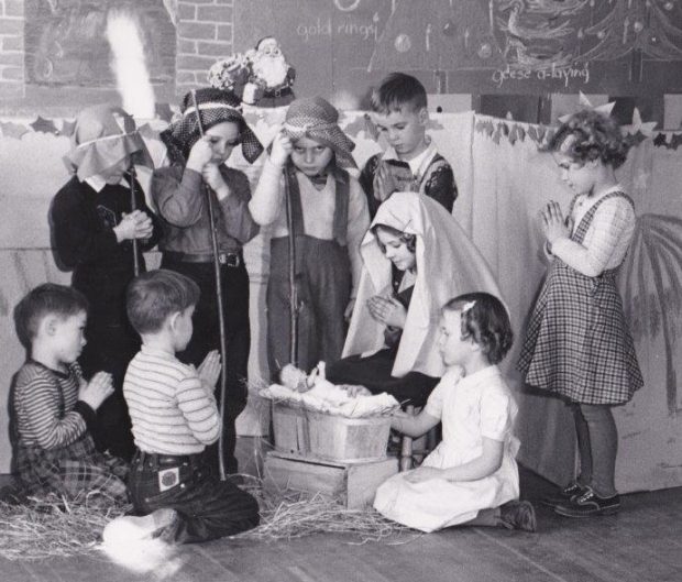 A group of young children are gathered in a Christmas nativity scene and most of the children have their hands held in prayer. They are looking at a doll in the manger.