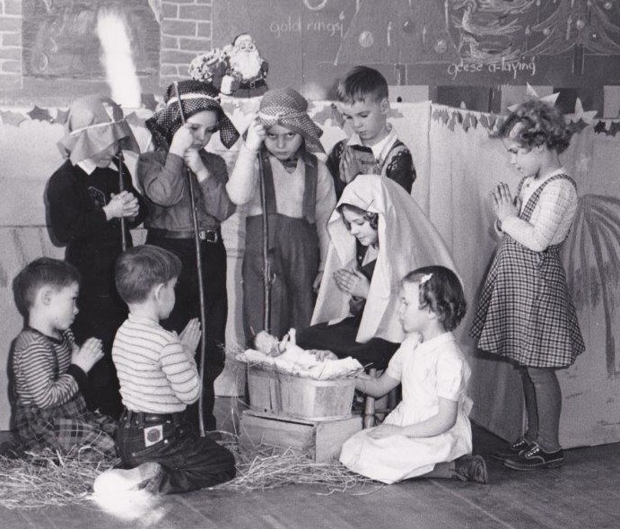 Un groupe d’enfants sont rassemblés à une crèche de Noël et plusieurs enfants tiennent leurs mains ensemble pour prier. Ils regardent une poupée dans la crèche.