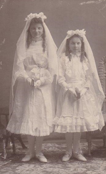 Two young girls are dressed in lacey dresses, gloves and a headdress with flowers. Both girls are holding a bible.