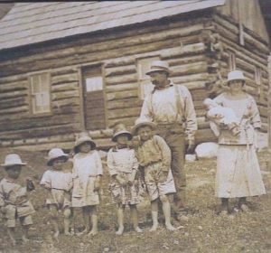A pioneer family is standing in front of a log cabin. The father is looking at the 5 small children standing in front of him. None of the children are wearing shoes and all are wearing hats. The mother is standing beside the man holding a baby.