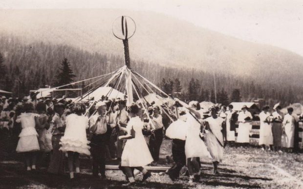 A group of children are dancing around the May pole. The girls are wearing frilly dresses and the boys are in dark pants and dress shirts. A group of pioneer are watching.