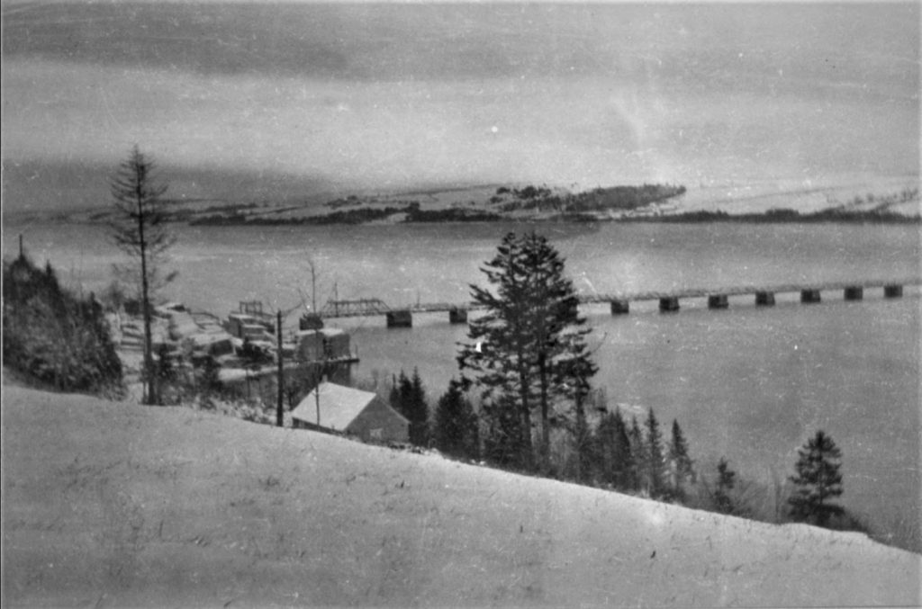 Long pont étroit traversant un large cours d’eau avec pont-levis près de la rive. Un peu de neige recouvre le sol et les toits des bâtiments. 