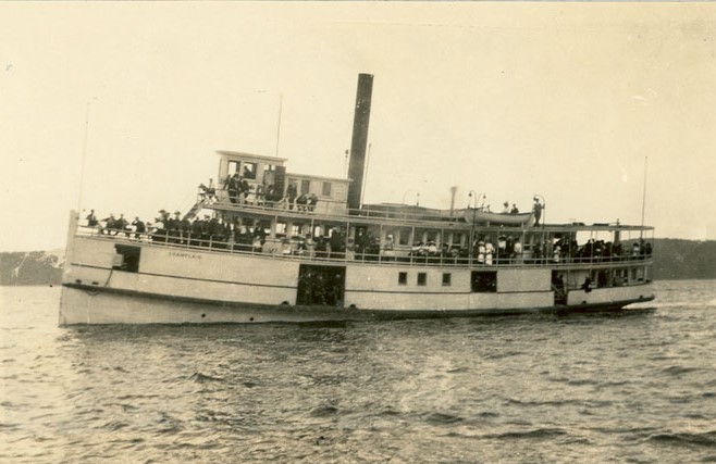 Port side view of a steam boat on the river. Passengers stand on the decks.