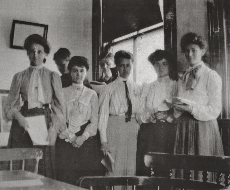 Five young women and two young men in period dress pose in a classroom.