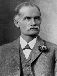 Older man facing slightly to his left. He has a moustache and is wearing a white shirt with a tweed vest and jacket.