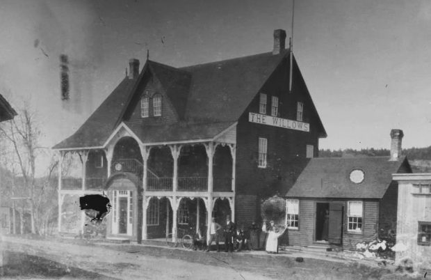 A three story building with an upstairs balcony. There is a one story wing on the right. A small group of men and women stand at the corner.