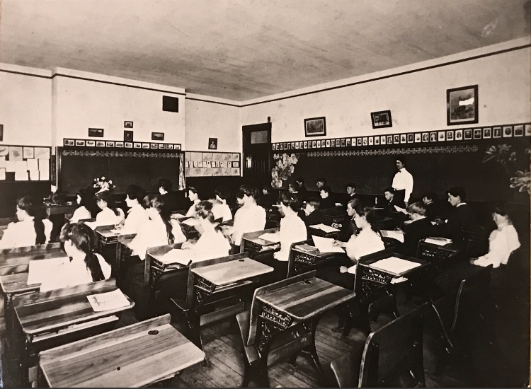 A female teacher stands at the blackboard to the right of her students who have their workbooks open on their desks.