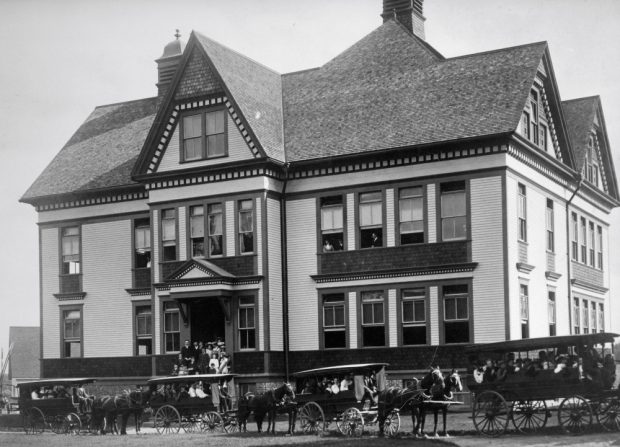 People stand on school steps. Others look out second storey windows. Students are in four school vans of varying sizes in front of the building. Each van has a driver and is led by two horses.